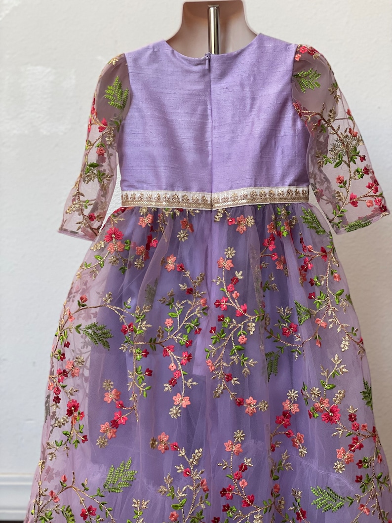 May include: A lavender dress with a floral embroidered tulle overlay. The dress has a round neckline and 3/4 sleeves. The bodice is made of a solid lavender fabric and the skirt is made of tulle with embroidered flowers in shades of pink, green, and gold.