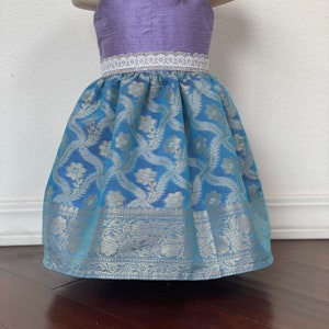 May include: A purple and blue sleeveless dress with a white lace trim around the waist. The skirt is made of a patterned fabric with silver accents.