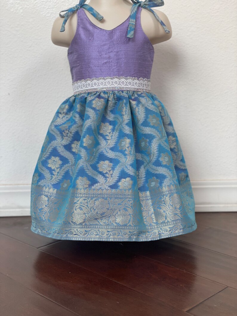 May include: A purple and blue sleeveless dress with a white lace trim around the waist. The skirt is made of a patterned fabric with silver accents.