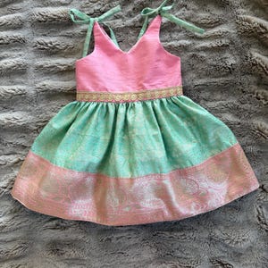 May include: A pink and mint green dress with a V-neck bodice and a gathered skirt. The bodice has tie straps, and the waist is accented with a gold trim. The skirt features a pink patterned border.