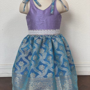 May include: A sleeveless dress with a lavender bodice and a blue skirt with a floral pattern. The dress has a white lace trim around the waist and blue ribbon ties at the shoulders.