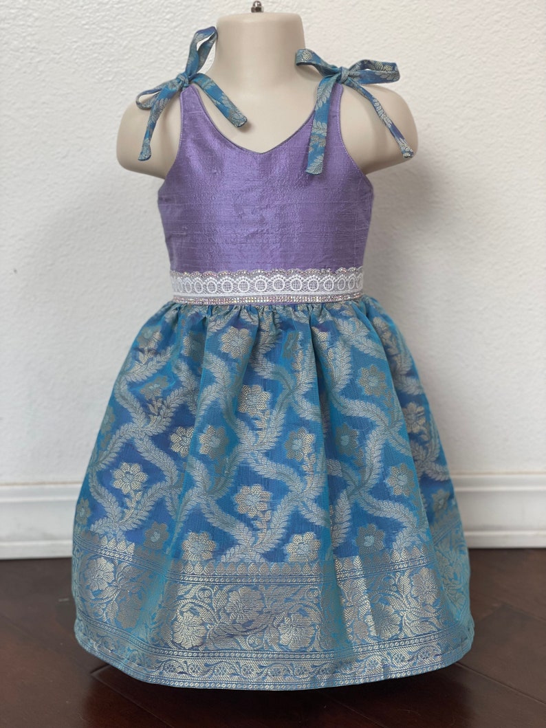 May include: A sleeveless dress with a lavender bodice and a blue skirt with a floral pattern. The dress has a white lace trim around the waist and blue ribbon ties at the shoulders.