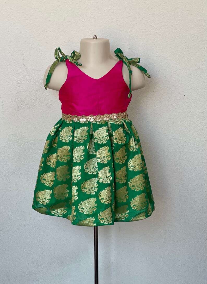 Puede incluir: Un vestido verde y rosa con ribete dorado y estampado de elefantes. El vestido tiene cuello en V y tirantes de lazo.