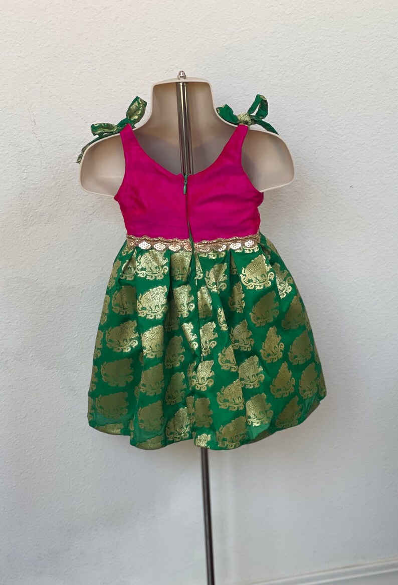 Puede incluir: Un vestido verde y rosa con detalles dorados y una cremallera en la espalda. El vestido tiene una falda verde con un estampado de elefantes dorados y un corpi&ntilde;o rosa con ribetes dorados. El vestido tiene tirantes verdes que se atan en lazos.