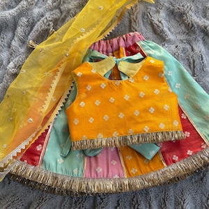 May include: A vibrant, traditional child's outfit. It includes a yellow top with a floral pattern and fringe, and a skirt in pink, red, and teal. A matching yellow scarf with silver detailing completes the ensemble.