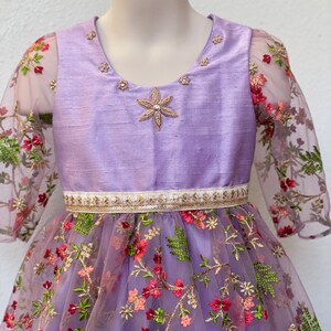 May include: A lavender dress with a floral embroidered tulle skirt and sheer sleeves. The bodice is a solid lavender color with a gold and white trim around the waist. The dress has a star-shaped embellishment on the bodice.