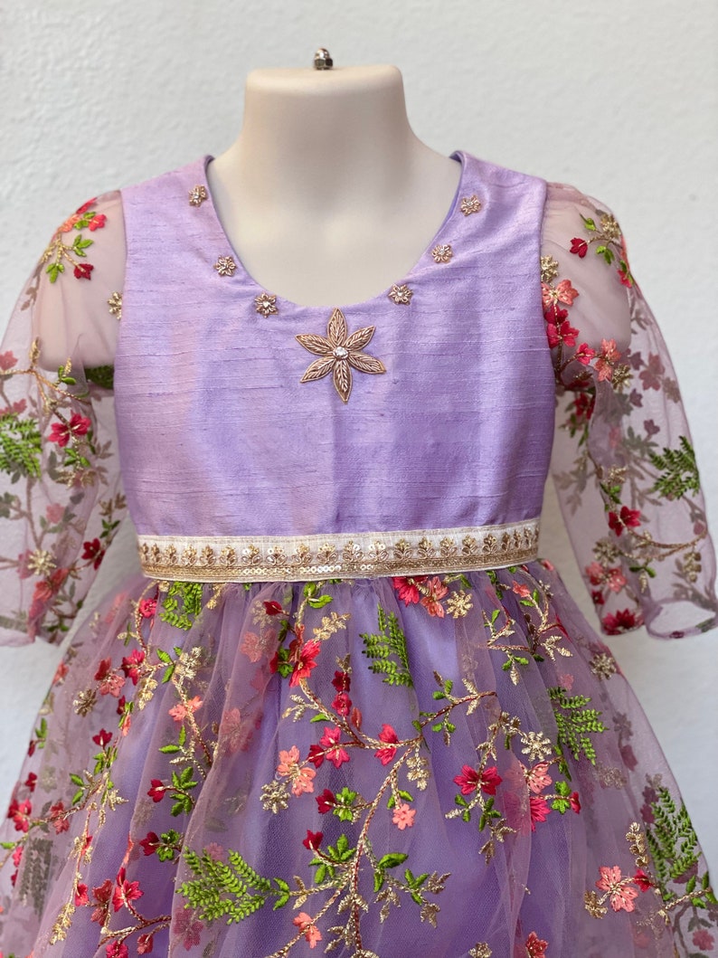 May include: A lavender dress with a floral embroidered tulle skirt and sheer sleeves. The bodice is a solid lavender color with a gold and white trim around the waist. The dress has a star-shaped embellishment on the bodice.