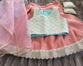 Op de afbeelding: Een traditionele Indiase outfit met een roze rok en een mintgroene top met ingewikkelde patronen. Een doorschijnende roze sjaal met gouden rand en kleine decoratieve kussens maken de set compleet. De outfit is voor een kind.