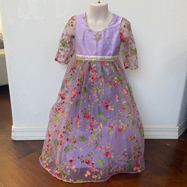 May include: A lavender dress with a floral embroidered tulle skirt and a solid lavender bodice. The dress has a white belt and three-quarter sleeves.