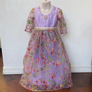 May include: A lavender-colored dress with a floral embroidered tulle overlay. The dress has a satin bodice with a silver star embellishment and a white satin belt.
