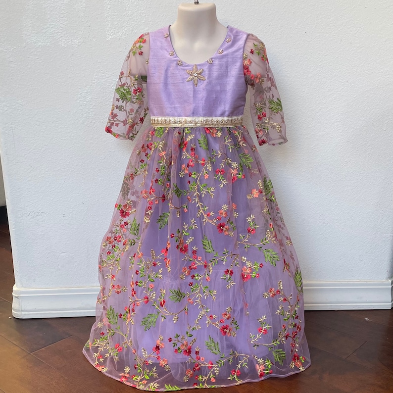 May include: A lavender-colored dress with a floral embroidered tulle overlay. The dress has a satin bodice with a silver star embellishment and a white satin belt.