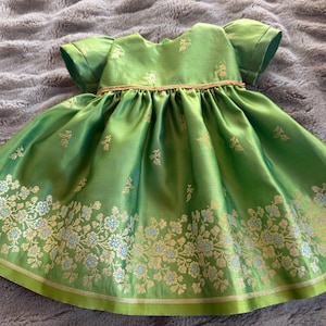 Puede incluir: Un vestido verde brillante con mangas cortas y falda amplia. El vestido presenta bordados florales dorados y celeste en el dobladillo y un ribete dorado en la cintura. La tela tiene un brillo sutil.