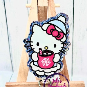 Puede incluir: Un charm Hello Kitty brillante azul y blanco con una taza roja y un diseño de copo de nieve. El charm está unido a un portagafete retráctil plateado con un charm de copo de nieve azul.