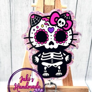 May include: A pink glitter resin keychain with a black and white sugar skull design. The skull has a pink bow and a spiderweb on the side. The keychain is attached to a retractable badge holder.