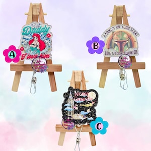 May include: Three decorative badge reels with glitter accents. One features Ariel with the text "But Daddy I love him", another has a Mandalorian helmet with "Karma's on your scent", and the last one shows Big Ben with "Never Grounded".