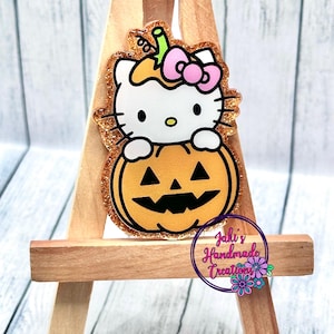May include: A white Hello Kitty character holding a glittery orange pumpkin with a jack-o'-lantern face. The pumpkin has a green stem and a pink bow on top. The character and pumpkin are on a wooden easel.
