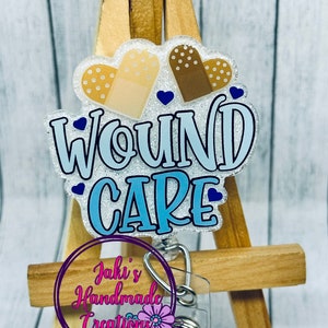 May include: A badge reel with the words "WOUND CARE" in blue and white, featuring band-aid heart shapes. The reel has a clear plastic clip and a metal retractable cord. The badge is displayed on a small wooden easel.