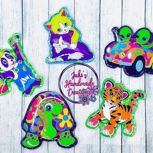 May include: Five colorful, glittery iron-on patches featuring a panda bear painting, a turtle with a psychedelic pattern, a tiger cub, two aliens in a car, and two cats. The patches are all on a white wooden background.