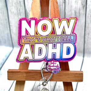 May include: A colorful badge reel with the text "NOW THAT'S WHAT I CALL ADHD" in a vibrant, eye-catching design. The badge is displayed on a small wooden easel, perfect for showcasing a unique accessory.
