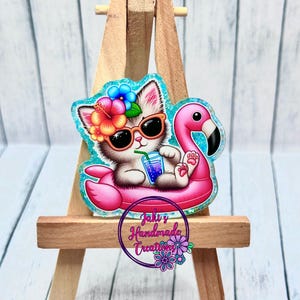 May include: A pink and white cat wearing sunglasses and a flower crown floats on a pink flamingo pool float. The cat is holding a blue and purple drink. The image has a blue and turquoise glitter background. The text "Jaki's Handmade Creations" is visible in the bottom right corner.
