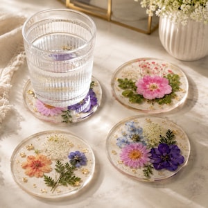 May include: Three round, clear resin coasters featuring embedded pressed flowers and gold flakes. One coaster supports a ribbed glass of water, while the others display arrangements of pink, blue, and white blossoms with green fern foliage.