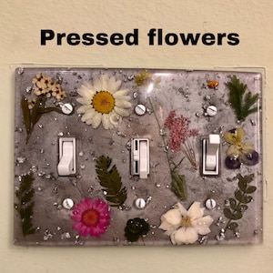 May include: A light switch cover with pressed flowers and silver glitter embedded in clear resin. The cover has three light switches, each with a white toggle.