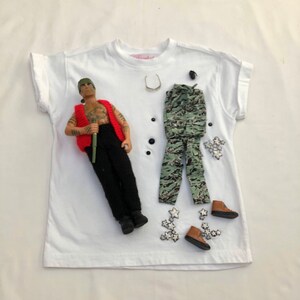 May include: A white t-shirt with a doll wearing a red vest, black trousers, and a green camouflage outfit. The doll is holding a stick. There are also several silver stars and a pair of black sunglasses on the shirt.