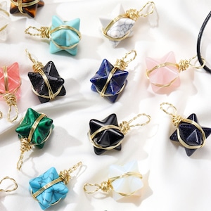 May include: A collection of star-shaped gemstone pendants in various colors, including turquoise, black, pink, and blue. Each pendant is wrapped with gold-colored wire and has a loop for hanging. The pendants are displayed on a white surface.