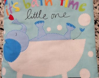 May include: A light blue baby bath book with the text "It's bath time little one." The book features an illustration of a blue elephant in a white bathtub with bubbles. The book is made of soft fabric.