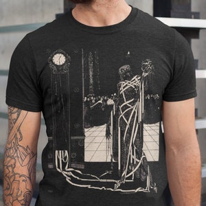 May include: Black t-shirt with a white graphic print of a woman tied up with ropes and a clock in the background. The graphic is a black and white illustration.