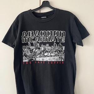 Peut inclure: Un t-shirt noir à manches courtes avec une impression graphique blanche représentant un groupe de créatures démoniaques à une table. Le texte ABADDON INCARNATE est imprimé en blanc au-dessus et THE LAST SUPPER est écrit en rouge en dessous.