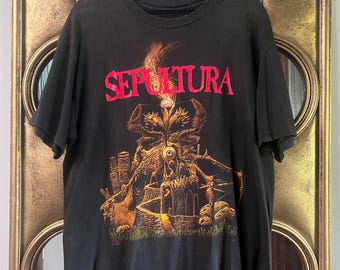 Puede incluir: Una camiseta negra de manga corta con un estampado gráfico que muestra la palabra SEPULTURA en letras rojas sobre una ilustración de un paisaje oscuro y surrealista con un ojo central, llamas y estructuras de piedra sobre hierba.