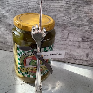 May include: A silver pickle fork with a middle finger design, tied with a black ribbon to a jar of pickles. The jar has a label that says "Tony's Pickles & Peppers, The Original". The tag on the ribbon says "Fickle Pickle Poker".