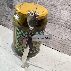 May include: A silver pickle fork shaped like a middle finger with a black ribbon tied around the handle. The fork is sticking out of a jar of green pickles with a yellow lid. The jar has a green and white checkered label that says "Tom's Pickled Peppers" and a white label that says "Fickle Pickle Poker".