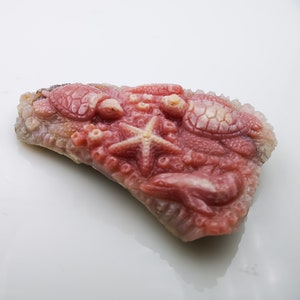 Puede incluir: Un colgante de piedra tallada en rosa y blanco con una estrella de mar, dos tortugas marinas y coral. El colgante está hecho de piedra pulida y tiene un acabado liso y brillante.
