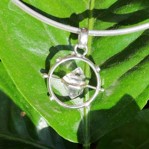 May include: A silver necklace with a clear crystal pendant. The crystal is set in a circular frame with a silver bar across the center. The necklace is on a green leaf.