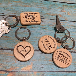May include: Wooden keyrings with engraved designs. One keyring has the text "Home isn't a place, 615, It's a feeling...". Other keyrings feature a heart with "37138", and "Home Sweet Home, 37122, Mt. Juliet, TN".