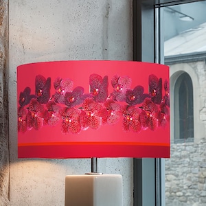 Może przedstawiać: Nowoczesna lampa stołowa z prostokątną, jasnoszarą podstawą i żywym czerwonym abażurem. Abażur zdobi kwiatowy wzór w odcieniach czerwieni i fioletu. Lampa stoi na parapecie.