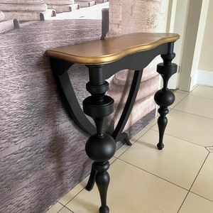 Puede incluir: Una mesa consola negra y dorada con una parte superior curva y patas ornamentadas. La mesa tiene una parte superior dorada y patas negras con remates decorativos de bola.