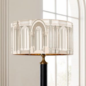 May include: A decorative lamp with a cylindrical lampshade featuring a classical architectural design in shades of white and beige. The lamp has a black base with gold accents, creating a sophisticated aesthetic. The lamp is a stylish home decor item.