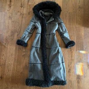 May include: A long, grey coat featuring a black faux fur hood, cuffs, and hem trim. The coat has a structured design with horizontal black piping details across the front, long sleeves, and a central button closure system along the front placket.