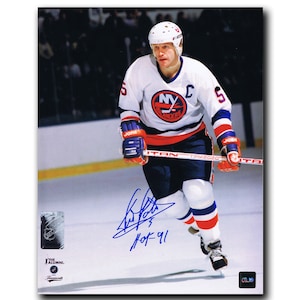 May include: A signed photo of a hockey player wearing a white jersey with the New York Islanders logo and the number 5. The player is skating on the ice and holding a hockey stick. The photo is signed "Mike Bossy" and "HOF-91".