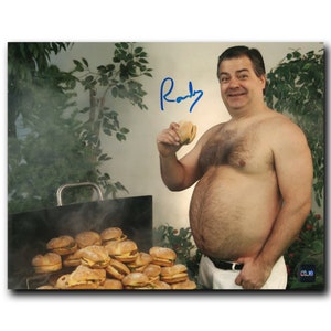 May include: A shirtless man with a large stomach holds a hamburger in one hand and smiles at the camera. He is standing in front of a grill with many hamburgers on it. The man's signature, "Randy", is written in blue ink in the top right corner of the image.