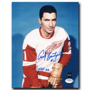 May include: A framed photograph of a man in a Detroit Red Wings hockey jersey. The jersey is white with red sleeves and accents, featuring the team logo and the number 1. The photo includes an autograph and a PSA authentication sticker.