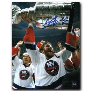 May include: A hockey player in a white jersey with a blue and orange New York Islanders logo raises a silver Stanley Cup trophy above his head. The player is surrounded by other players and fans. The image is a black and white photograph.