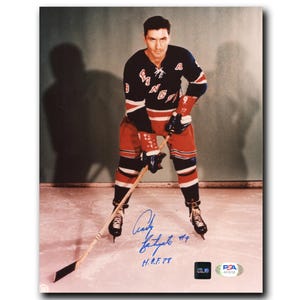 May include: A framed photograph of a hockey player in a New York Rangers uniform, holding a hockey stick. The player is wearing a navy blue jersey with red and white accents, red pants, and black skates. The photo includes the player's signature and the text "H.O.F. 78".