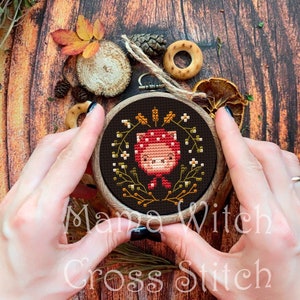 May include: A cross stitch pattern of a cute cartoon mushroom with a red cap and white spots, surrounded by a wreath of green leaves and white flowers. The pattern is framed in a wooden hoop and held by a person's hands.