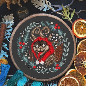 May include: A cross stitch pattern of a cute, furry creature wearing a red hooded hat, surrounded by a wreath of green leaves and red berries. The pattern is framed in a wooden hoop and displayed on a black background.