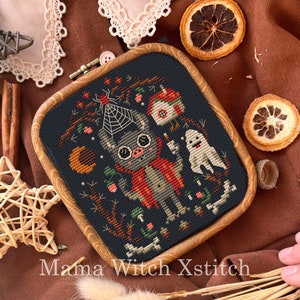 May include: A cross-stitch embroidery in a wooden frame depicts a cute cartoon monster wearing a red cape and holding a candle. The monster is standing next to a ghost and a small house. The background is a dark blue with a moon, stars, and other Halloween-themed elements.