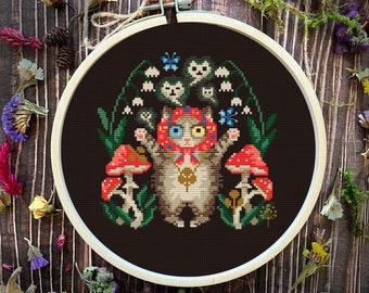 Op de afbeelding: Een borduurring met kruissteek met een zwarte achtergrond waarop een kat met een rode hoed en een paddenstoel in zijn hand is afgebeeld. De kat heeft één blauw oog en één geel oog. De kat is omringd door paddenstoelen, bloemen en vlinders.